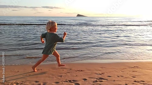 Blonde 3-years-old girl in knitted sweater laughing runs along water's edge on city's sandy beach at sunset. Long shot. Slow motion.