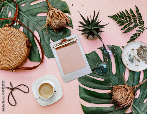 Flat lay women's office desk. Female workspace with clip board, tropical leaves, accessories, cup of coffee on pink background. Top view .Copy space photo