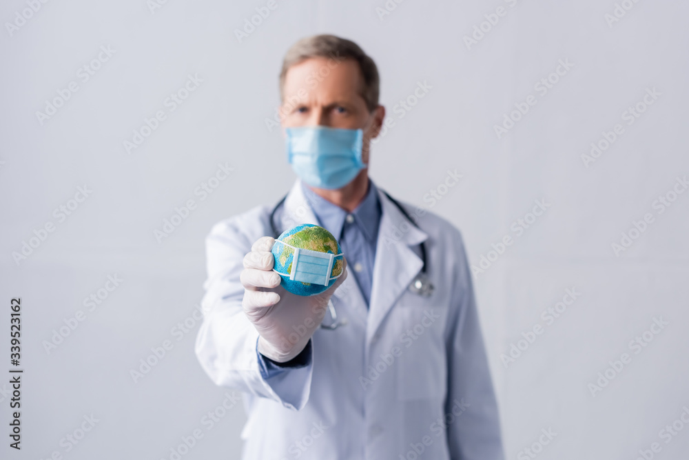 selective focus of middle aged doctor in latex glove and medical mask holding small globe on grey