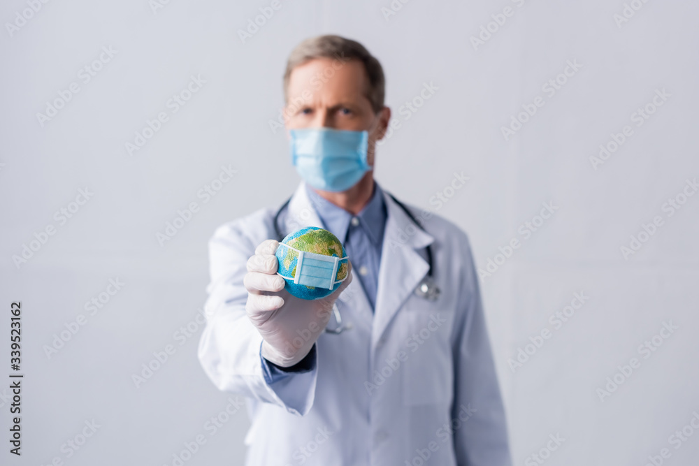 selective focus of middle aged doctor in latex glove and medical mask holding small globe on grey selective focus of middle aged doctor in latex glove and medical mask holding small globe on grey