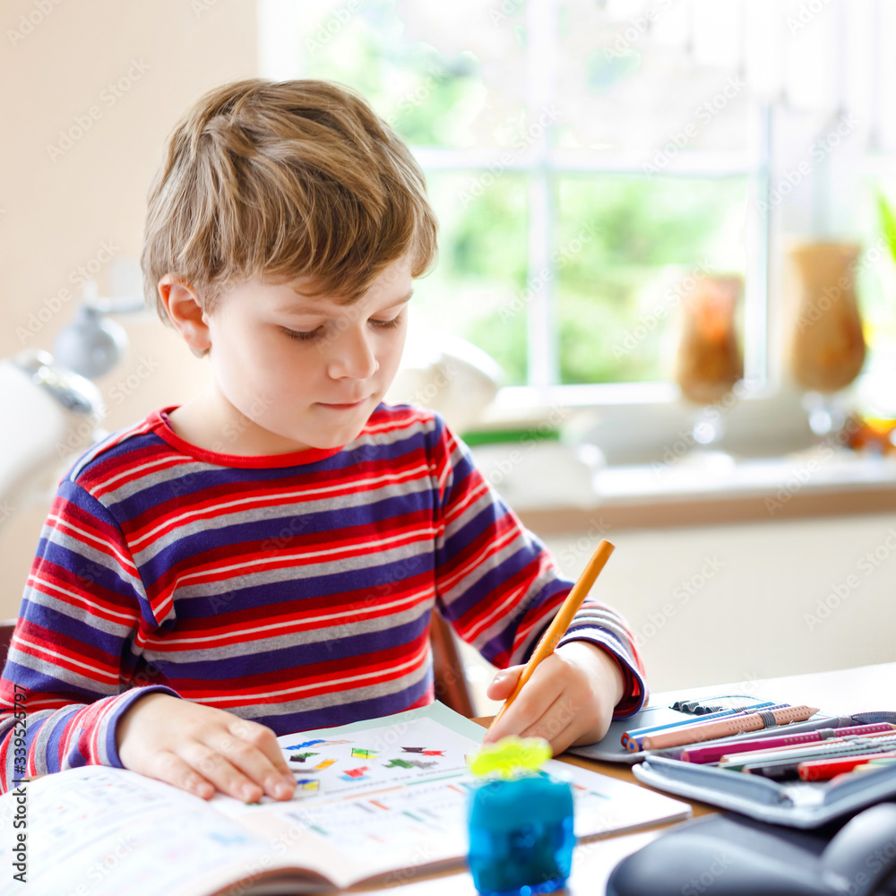 Hard-working happy school kid boy making homework during quarantine time from corona pandemic disease. Healthy child writing with pen, staying at home. Homeschooling concept