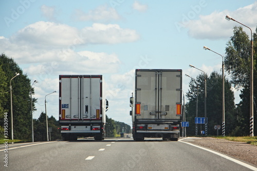 Two parallel course semi trucks on summer road on trees and streetlightl on roadsides and blue sky with clouds background