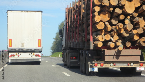 Two vehicles with semi trailers - van and wood truck on the road in summer day on blue sky background, safety overtaking driving, fast transportation logistics delivery on suburban asphalted highway