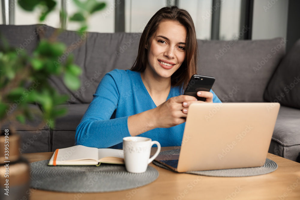 Image of smiling young cute woman using laptop and mobile phone Image of smiling young cute woman using laptop and mobile phone