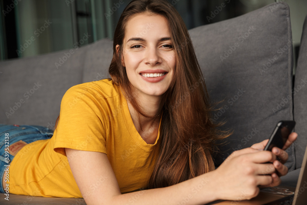 Image of smiling woman using laptop and mobile phone while lying Image of smiling woman using laptop and mobile phone while lying