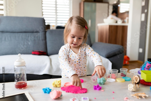 Happy little child, adorable creative 2 year old girl playing with dough, colorful modeling compound, sitting bright sunny room at home.