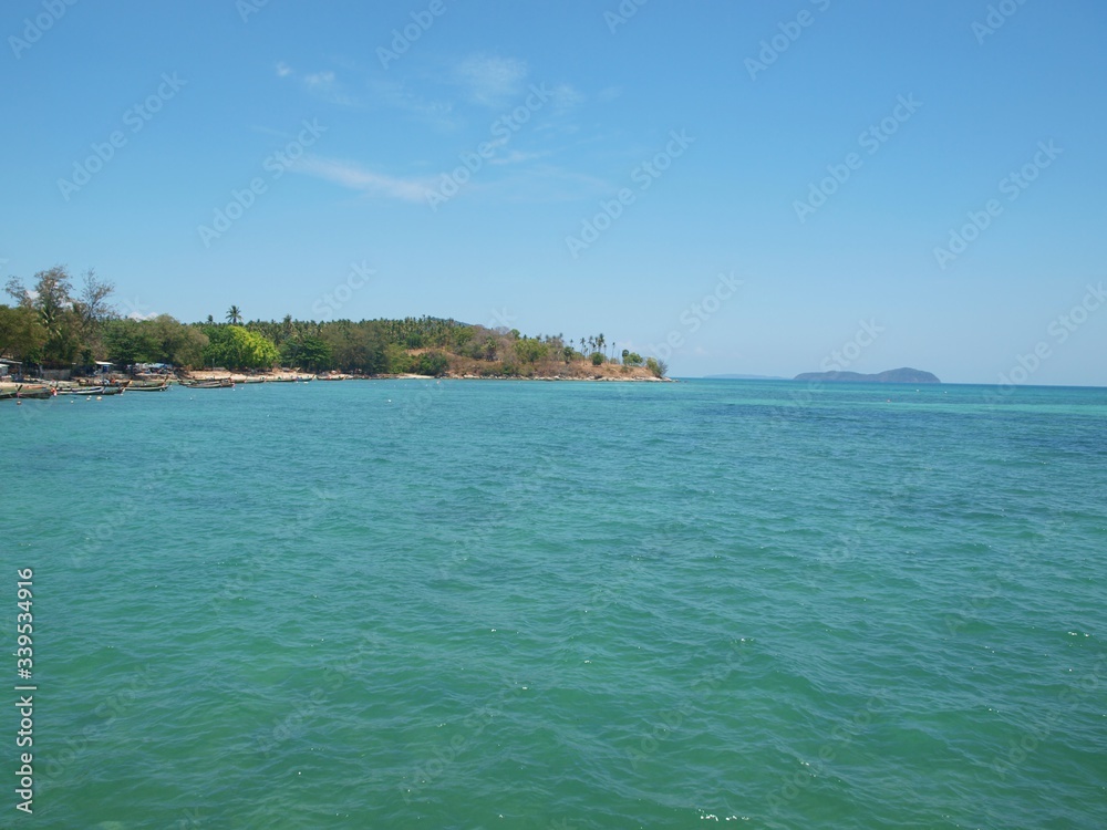 Panoramic view to the sea, green cliff covered by rainforest, small island on the horizon, azure color calm water and sandy coast with longtailboats on it. Seascape and tropical nature. Blue sky. 
