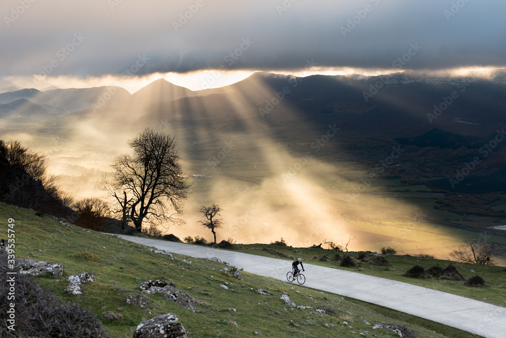 Fototapeta premium A romantic cyclist climbing a lonely road while the sunrise illuminates with beautiful light the valley, San Miguel de Aralar, Navarra, Spain.