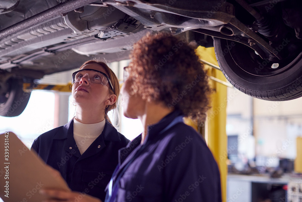 Female Tutor With Student Looking Underneath Car On Hydraulic Ramp On Auto Mechanic Course 