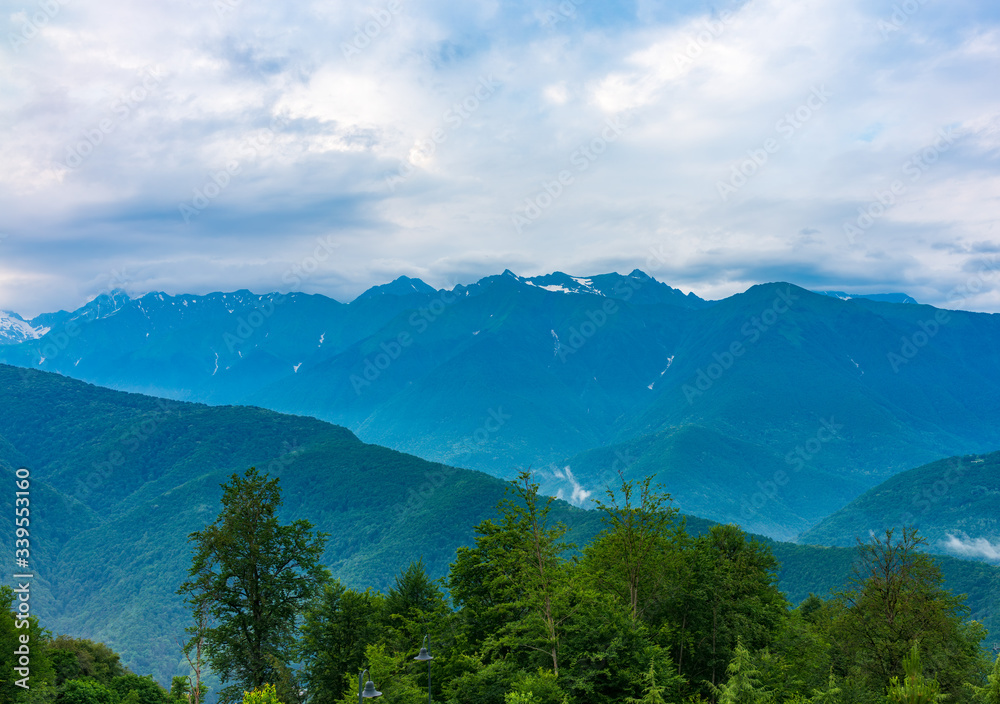 Green mountain ridges, surrounded by high mountains. Snow-capped mountain peaks on the horizon. Mountain landscape with thick clouds. Krasnaya Polyana, Sochi, Caucasus, Russia. Green mountain ridges, surrounded by high mountains. Snow-capped mountain peaks on the horizon. Mountain landscape with thick clouds. Krasnaya Polyana, Sochi, Caucasus, Russia.