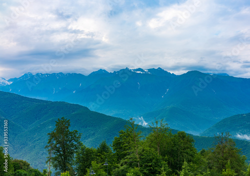 Green mountain ridges, surrounded by high mountains. Snow-capped mountain peaks on the horizon. Mountain landscape with thick clouds. Krasnaya Polyana, Sochi, Caucasus, Russia. © Dmitrii Potashkin