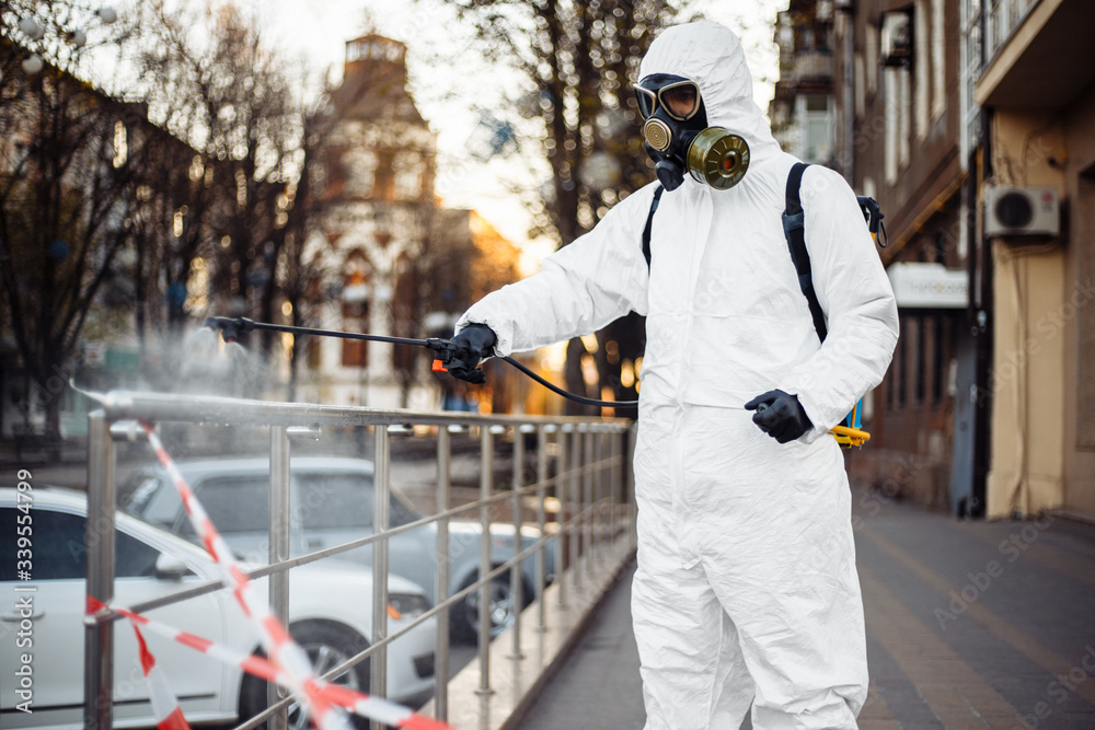 A man in protective equipment disinfects with a sprayer in the city. Surface treatment due to coronavirus covid-19 disease. A man in a white suit disinfects the street and rails. Virus pandemic.