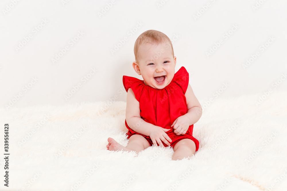an eight-month-old cute Caucasian baby girl in a red suit sits on the bed and laughs. an eight-month-old cute Caucasian baby girl in a red suit sits on the bed and laughs.