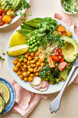 Healthy vegetarian lunch bowl with avocado, chickpeas, quinoa and vegetables, garnished with microgreens and nut dressing. Flat lay on stone background.