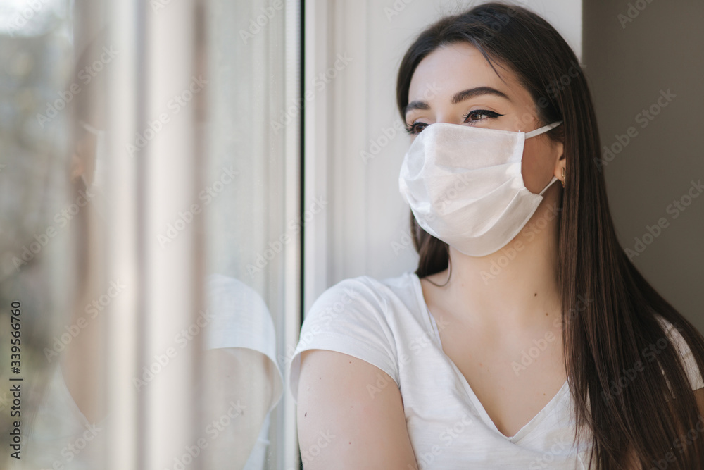 Young woman sits on windowsill in protective mask and look out the window. Alone at home in quarantine. Coronavirus theme. Stay at home Young woman sits on windowsill in protective mask and look out the window. Alone at home in quarantine. Coronavirus theme. Stay at home