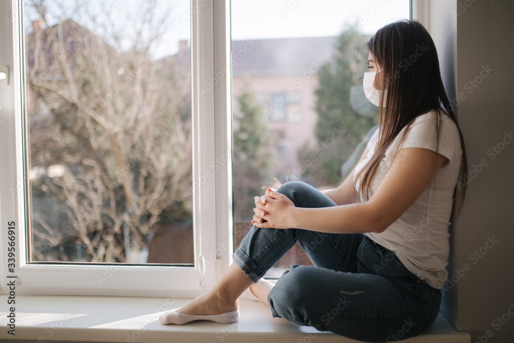 Side view of Young woman sits on windowsill and in protective mask and look out the window. Alone at home in quarantine. Coronavirus theme. Stay at home Side view of Young woman sits on windowsill and in protective mask and look out the window. Alone at home in quarantine. Coronavirus theme. Stay at home