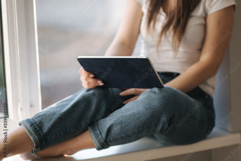 Mid selection of Attractive young woman in white medical mask sits on windowsiil and use tablet. Female have a rest at home in quarantine Mid selection of Attractive young woman in white medical mask sits on windowsiil and use tablet. Female have a rest at home in quarantine