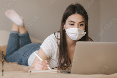 Young woman in mask use laptop on the bed. Female working during quarantine in bedroom. Student study at home. Stay at home
