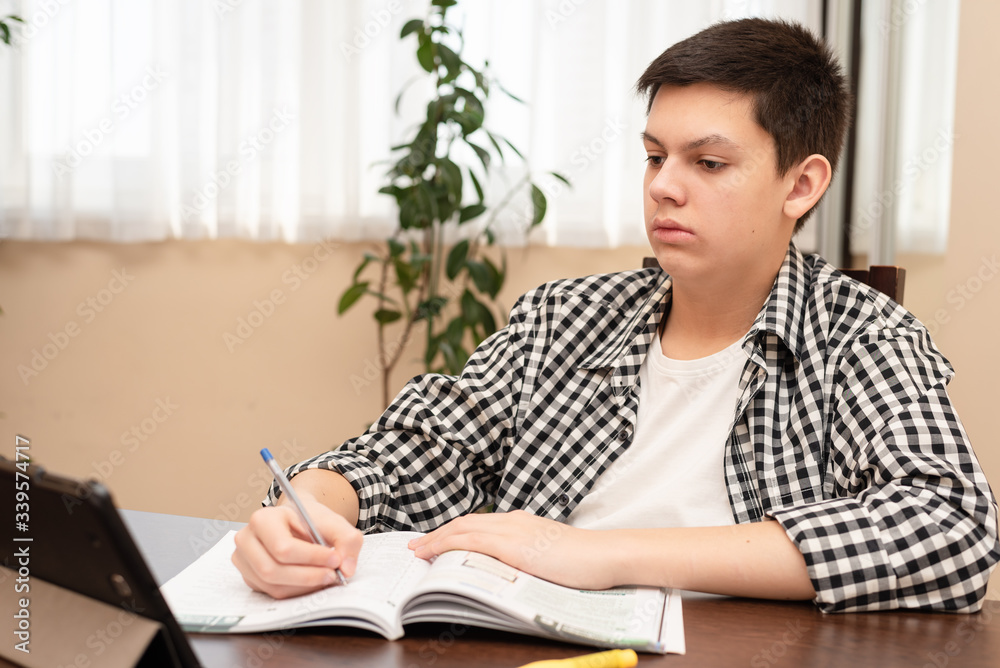 Teenage boy on distance learning at home. Student studying at home with digital tablet doing school homework in training notebook. Distance online education consept. Staying at home during quarantine. Teenage boy on distance learning at home. Student studying at home with digital tablet doing school homework in training notebook. Distance online education consept. Staying at home during quarantine.