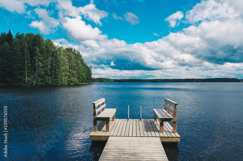 Fototapeta Naklejka Na Ścianę i Meble -  Two wooden chairs bench on a wood pier overlooking a blue lake water with green forest and cloud sky