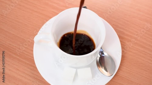 Slow motion coffee pour into a white porcelain cup on wooden table. Sugar cubes and metal spoon on the small plate.
