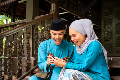 Young couple of malay muslim in traditional costume watching online content in a smart phone with happy expression during Eid al-Fitr celebration.