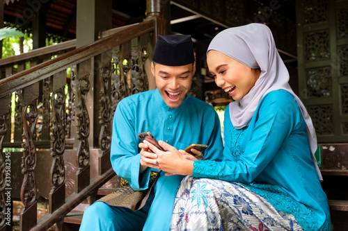 Young couple of malay muslim in traditional costume watching online content in a smart phone with happy expression during Eid al-Fitr celebration.