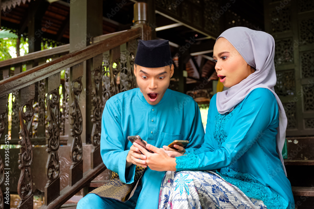Young couple of malay muslim in traditional costume watching online content in a smart phone with surprise expression during Eid al-Fitr celebration.
