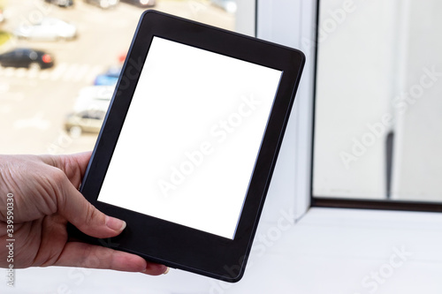 A modern black electronic book with a white blank screen in female hand against a blurred window background. Mockup tablet closeup