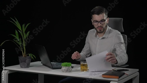 Caucasian attractive bearded male copywriter in glasses typing text on laptop and and looking to paper sheets at white desk. Indoor studio shot on black background.