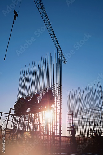 The construction of a rooftop high rise building, workers are tied with steel bars for concrete pouring, surveying the site and cranes are lifting or moving metal at the construction site. Pastel tone