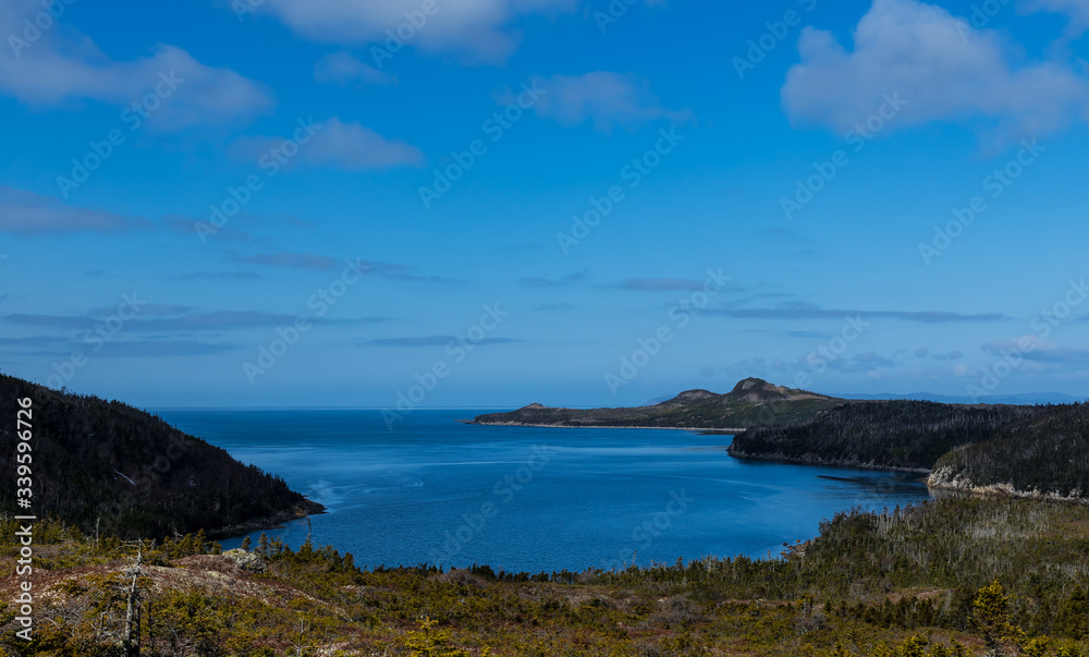 A small cove with a deep blue ocean, blue skies, clouds and rocky land. The circular shape of the bay has rocky cliffs, forest and sandy beaches. The water is calm and the sky has some light clouds. 