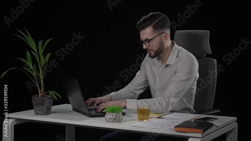 Tired of working on laptop young bearded man in a stylish look reclining on chairs back and puts arms behind his head. Indoor studio shot on black background.