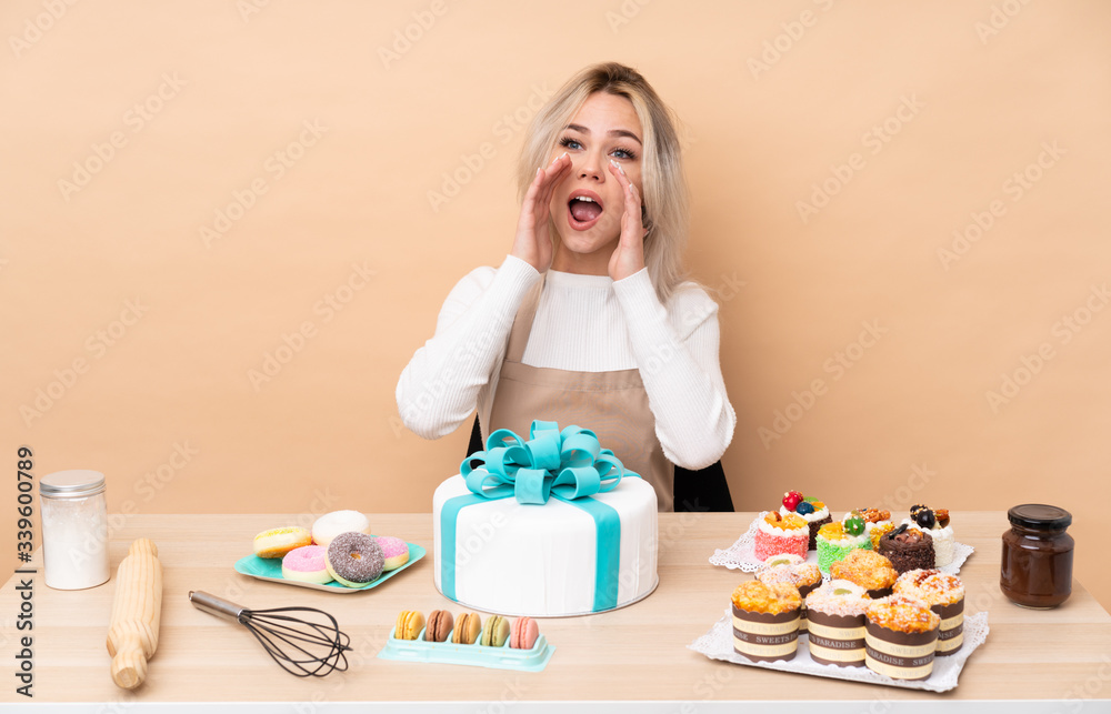 Teenager pastry chef with a big cake in a table shouting with mouth wide open
