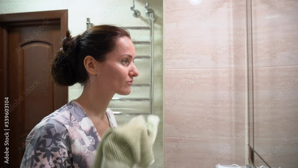Beautiful cute Caucasian woman washed her face, rubs with a green towel, wearing protective medical mask, looks at herself in front of the mirror in bathroom. Covid 19 virus protection concept. Beautiful cute Caucasian woman washed her face, rubs with a green towel, wearing protective medical mask, looks at herself in front of the mirror in bathroom. Covid 19 virus protection concept.