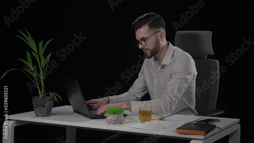 Tired of working on laptop young bearded man in a stylish look puts off his glasses and rubbs his eyes. Indoor studio shot on black background.