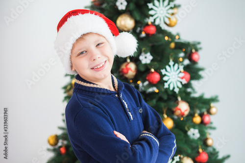 Closeup portrait of little white kid standing in Christmas holiday interior near big decorated green tree smiling happily and looking at camera. Boy wearing red Santa hat.