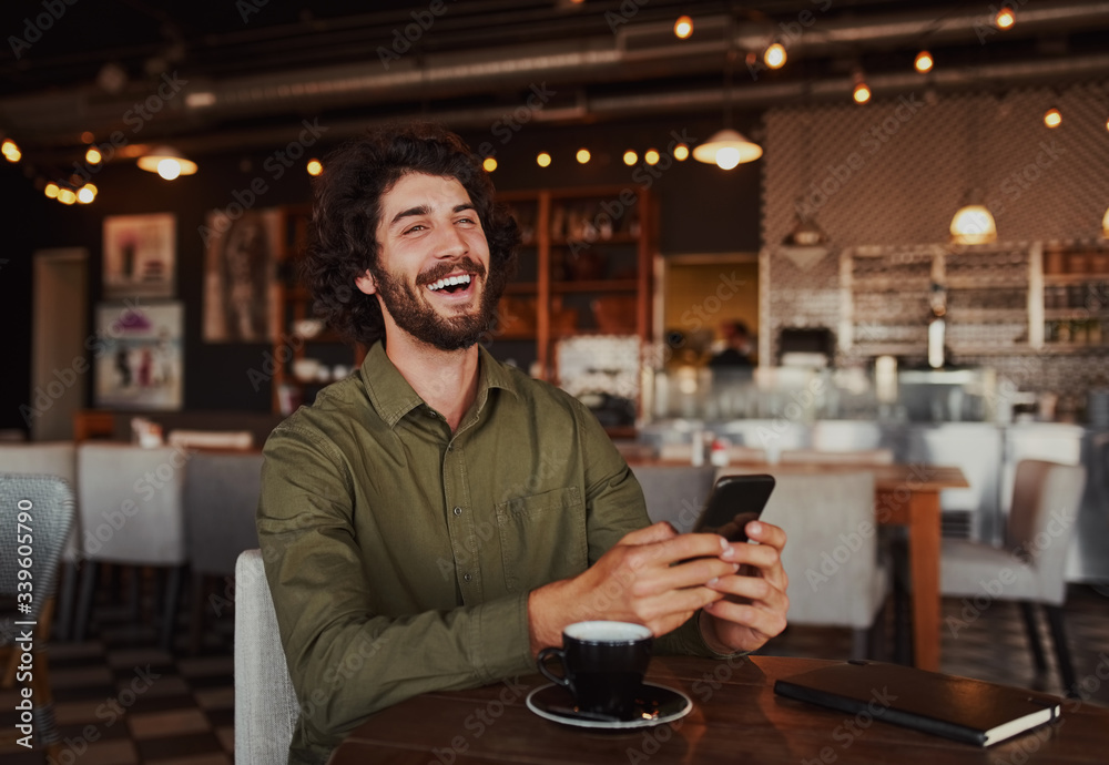Portrait of handsome young man laughing while reading funny message on phone sitting in cafeteria during free time