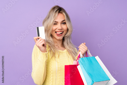 Teenager blonde girl over isolated purple background holding shopping bags and a credit card