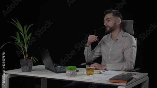 Young attractive bearded male copywriter looking for idea rotating in chair puts on his glasses and starts typing. Indoor studio shot on black background.