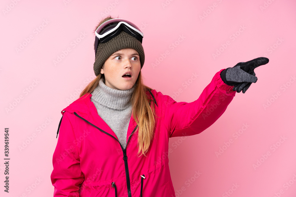 Ukrainian teenager skier girl with snowboarding glasses over isolated pink background pointing away