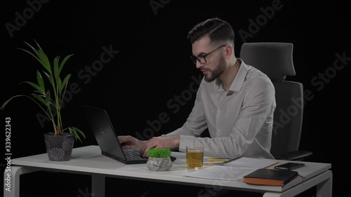 Bearded young businessman is talking on mobile phone during working on laptop. Business communication, modern technology and millennials concept. Indoor studio shot on black background.