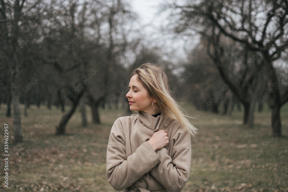 Young beautiful woman in a beige coat walks alone in a park on the street. Resting, windy weather, positive emotions, power balance. Stylish clothes for autumn, spring. Young beautiful woman in a beige coat walks alone in a park on the street. Resting, windy weather, positive emotions, power balance. Stylish clothes for autumn, spring.