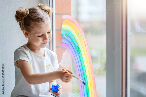 A little girl with blond hair draws a rainbow on the glass with paints. 