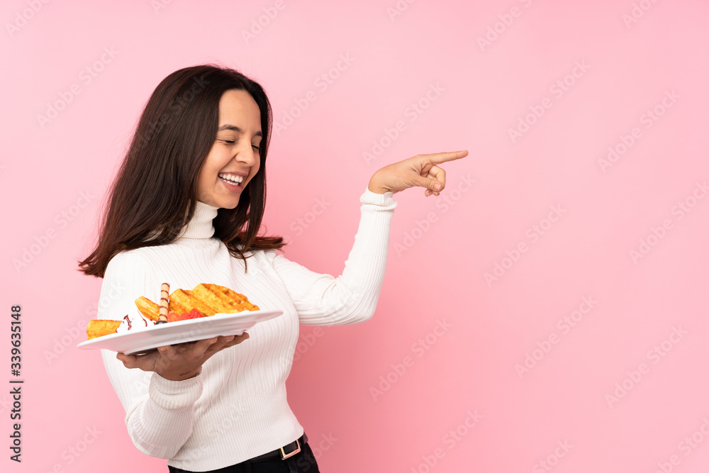 Young brunette woman holding waffles over isolated pink background pointing finger to the side and presenting a product Young brunette woman holding waffles over isolated pink background pointing finger to the side and presenting a product