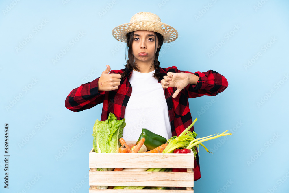 Young farmer Woman holding fresh vegetables in a wooden basket making good-bad sign. Undecided between yes or not