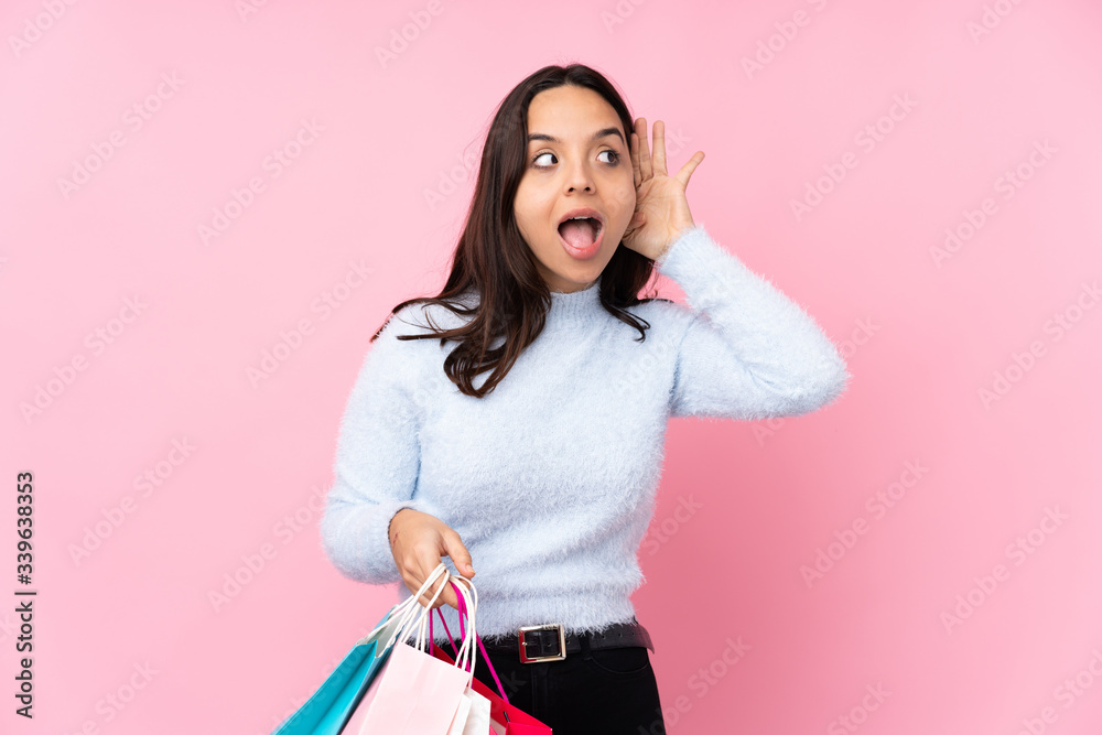 Young woman with shopping bag over isolated pink background listening to something by putting hand on the ear