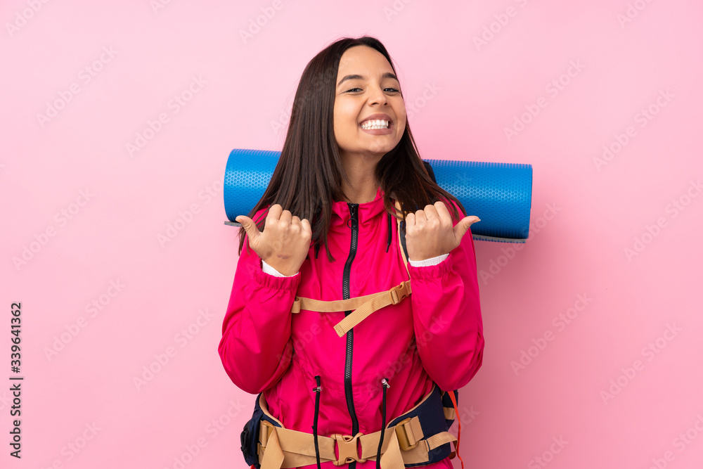 Young mountaineer girl with a big backpack over isolated pink background with thumbs up gesture and smiling Young mountaineer girl with a big backpack over isolated pink background with thumbs up gesture and smiling