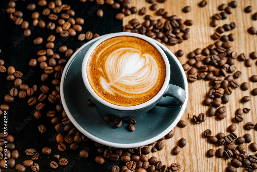 Obraz premium Gray cup of coffee on the black and brown table among spilled roasted coffee beans. Cappuccino with cofee art in the cafe.
