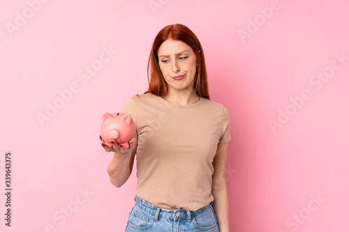 Young redhead woman over isolated pink background holding a big piggybank © luismolinero
